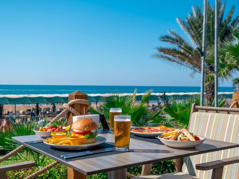 Mesa con hamburguesa, papas fritas y bebidas en terraza con vista al mar y palmeras al fondo