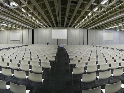 Grote conferentieruimte met meerdere rijen witte stoelen en tafels onder plafondverlichting.