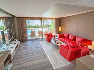 Ruime hotelkamer met rood meubilair, glazen tafel, tv en balkon met zeezicht.