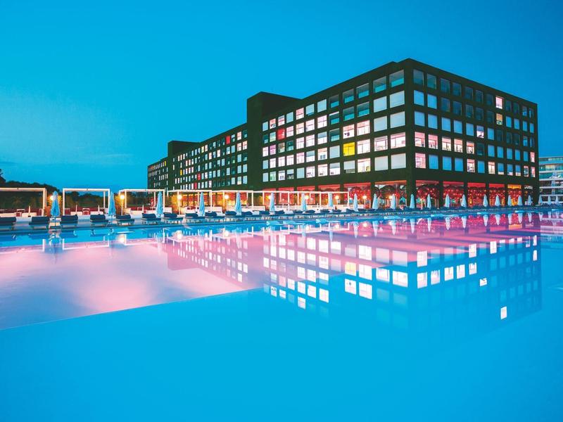 Modern hotel met verlichte ramen naast een groot, verlicht zwembad bij schemerlicht.