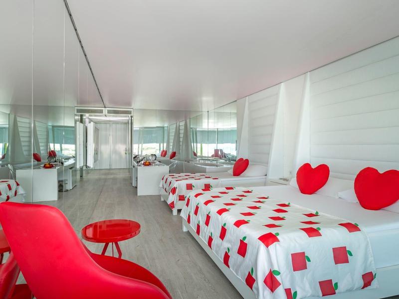 Lichte hotelkamer met twee bedden, rode hartkussens en een rode stoel en tafel.