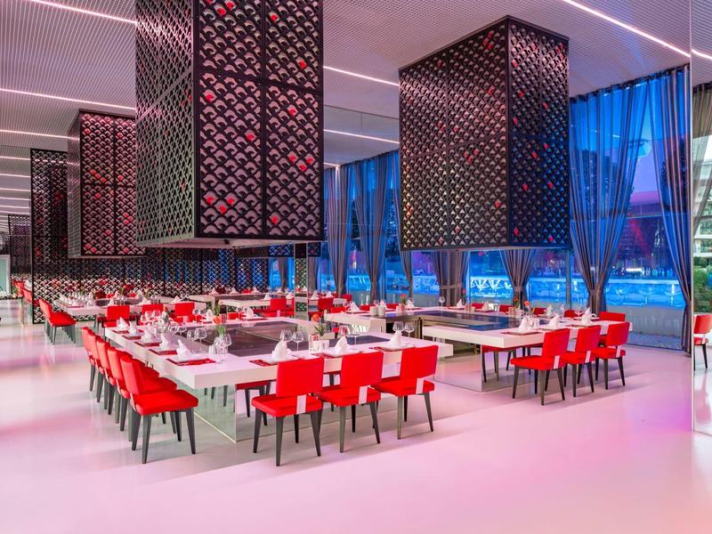 Modern restaurant met rode stoelen, grote ramen en geometrische plafondlampen in een ruime omgeving.