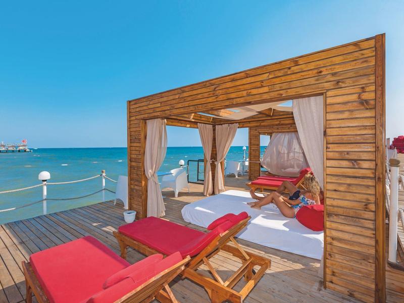 Houten cabana met rode ligstoelen en witte gordijnen op pier met uitzicht op zee.