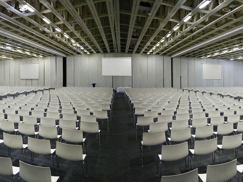 Grote conferentieruimte met meerdere rijen witte stoelen en tafels onder plafondverlichting.