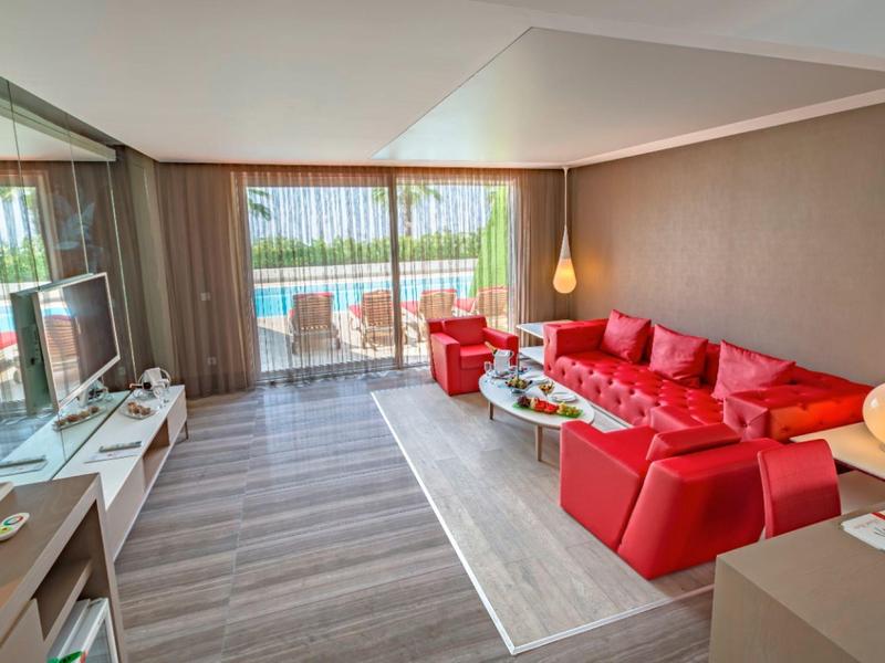 Ruime hotelkamer met rood meubilair, glazen tafel, tv en balkon met zeezicht.