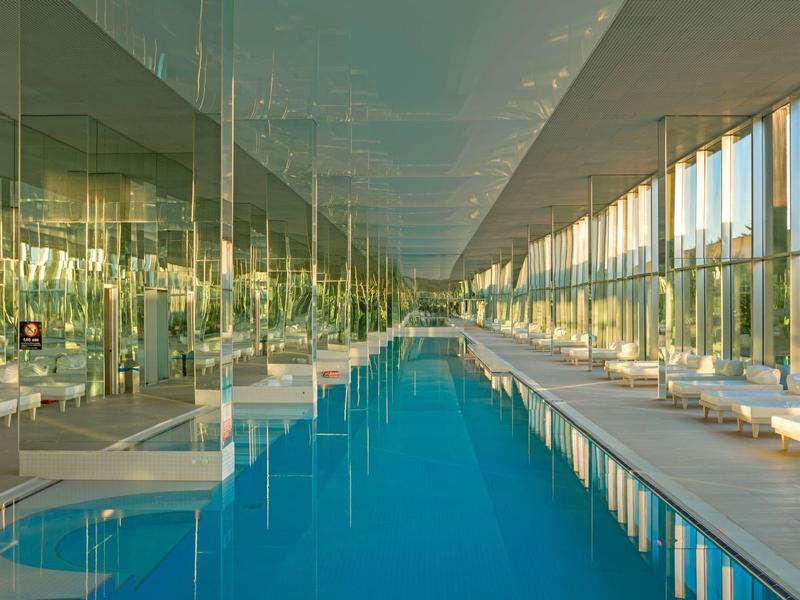 Moderne binnenzwembad met glazen wanden en comfortabele ligstoelen rechts.