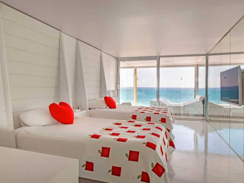 Modern hotelkamer met twee bedden, rode accenten en uitzicht op zee door grote ramen.