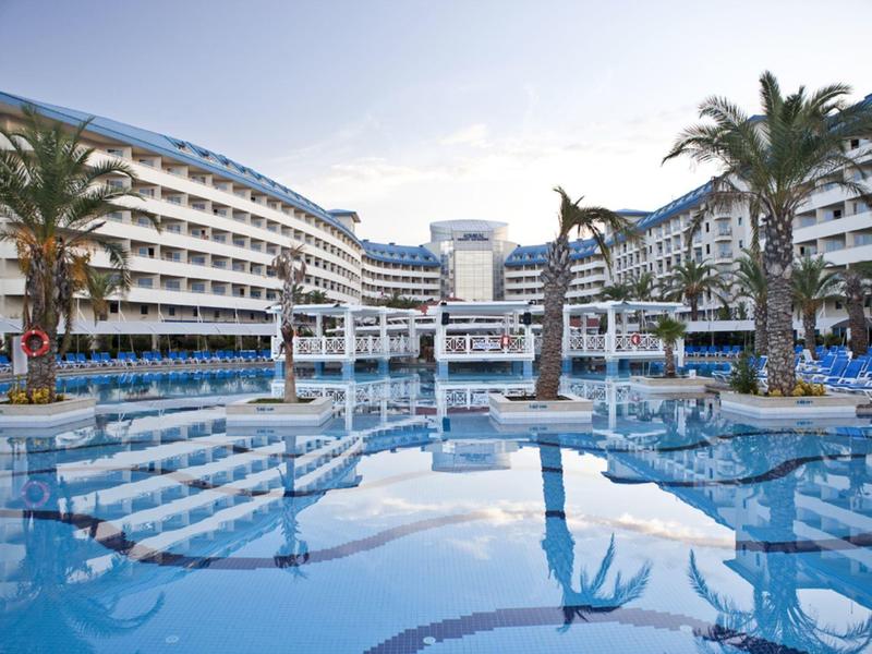 Grand bassin d'hôtel avec palmiers et chaises longues devant un bâtiment hôtelier à plusieurs étages sous un ciel clair.