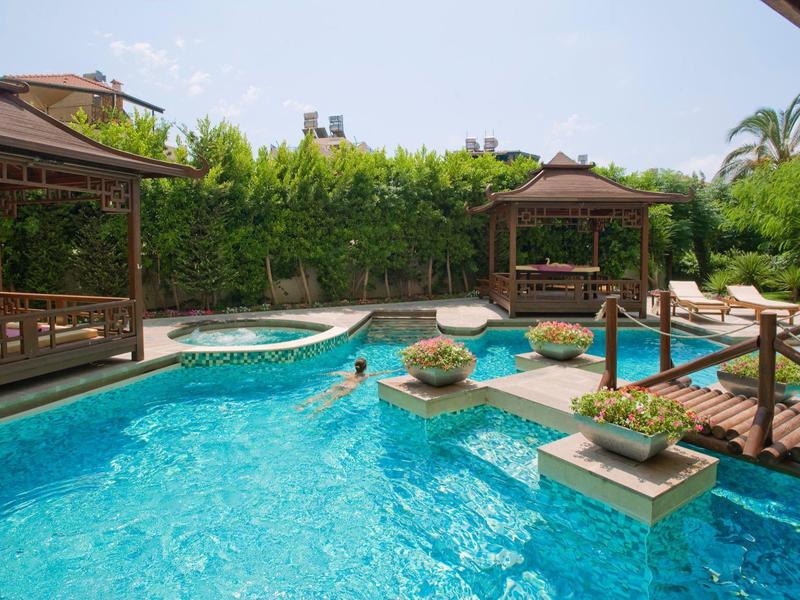 Piscine avec pavillons, plantes et un petit pont sous un ciel clair dans le jardin de l'hôtel.