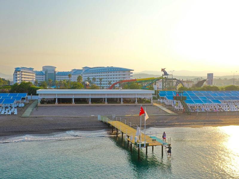 Une longue jetée avec un drapeau rouge et des personnes, avec une plage et des bâtiments d'hôtel modernes au coucher du soleil en arrière-plan.