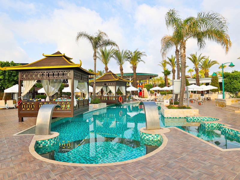 Espace piscine avec pavillon, palmiers et chaises longues sous un ciel bleu dans une station.