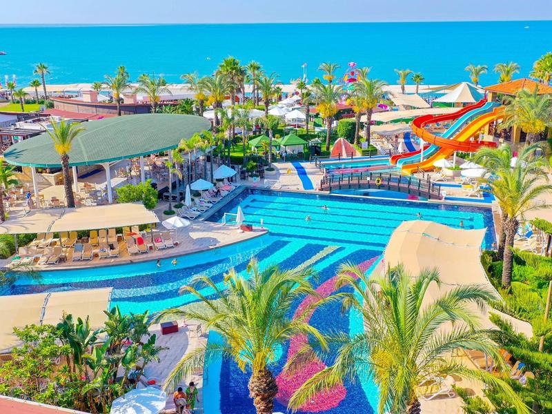 Complexe coloré avec piscine, toboggans aquatiques, palmiers et vue sur la mer sous un ciel clair.