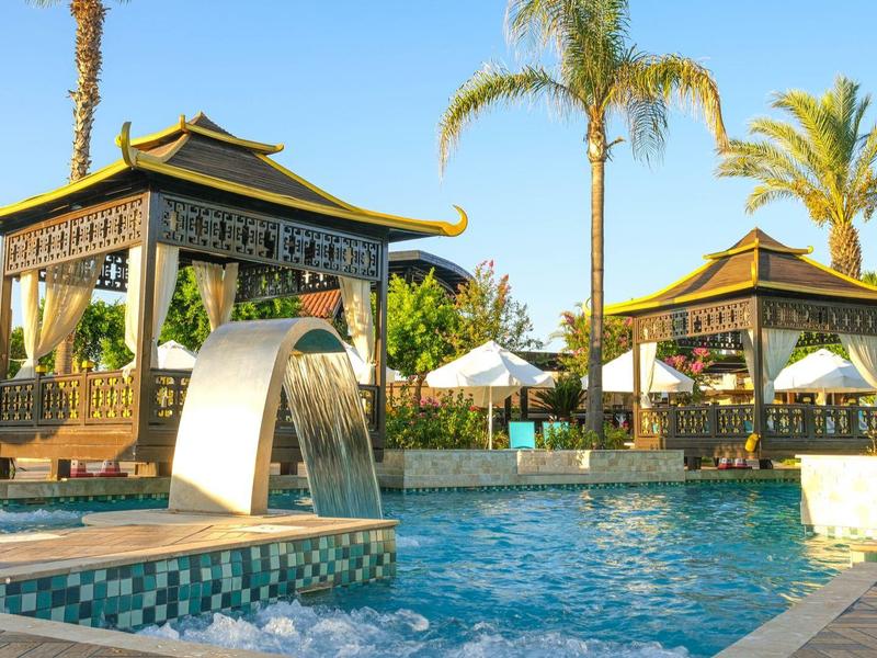 Piscine d'hôtel avec cascade, palmiers tropicaux et parasols sous des pavillons.