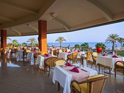 Offenes Restaurant mit gedeckten Tischen, Stühlen, Blick auf Palmen und das blaue Meer im Hintergrund.