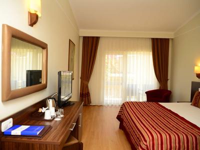 Hell eingerichtetes Hotelzimmer mit Doppelbett, Fernseher auf Holzkommode und großem Fenster mit Vorhängen.