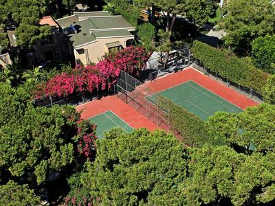 Widok z lotu ptaka na kort tenisowy otoczony drzewami i budynkami.