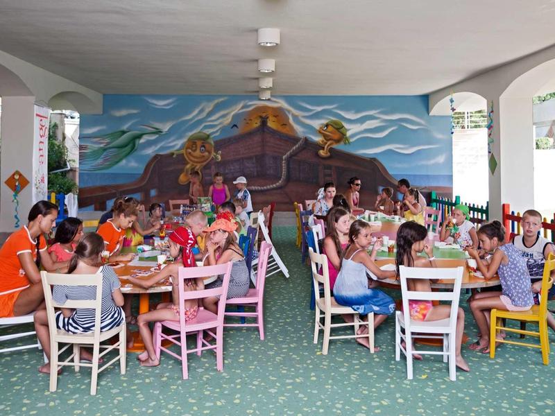 Kinder sitzen an bunten Tischen und essen gemeinsam in einem Raum mit großer Wandmalerei.