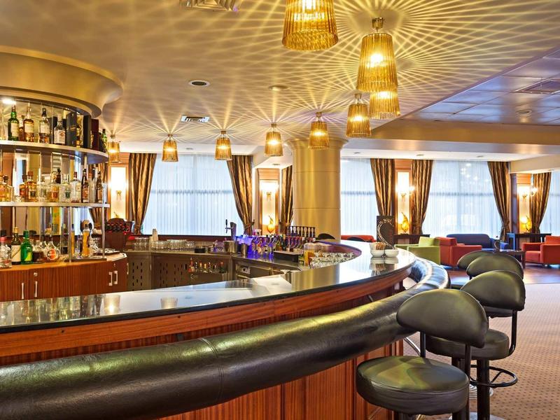 Elegante Bar mit rundem Tresen, gepolsterten Barhockern, goldfarbenen Lampen und großen Fenstern.