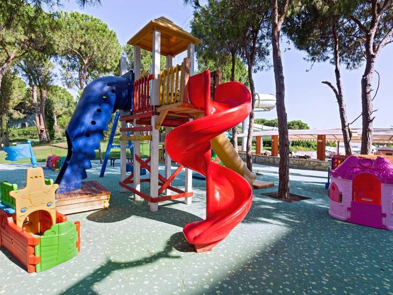 Bunter Spielplatz mit Rutsche, Klettergerüst und kleinen Spielhäusern unter Bäumen.