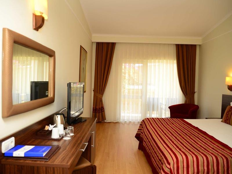 Hell eingerichtetes Hotelzimmer mit Doppelbett, Fernseher auf Holzkommode und großem Fenster mit Vorhängen.