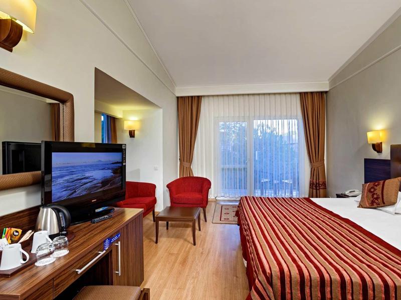 Hell eingerichtetes Hotelzimmer mit Doppelbett, rotem Teppich, zwei Sesseln und großem Fenster.