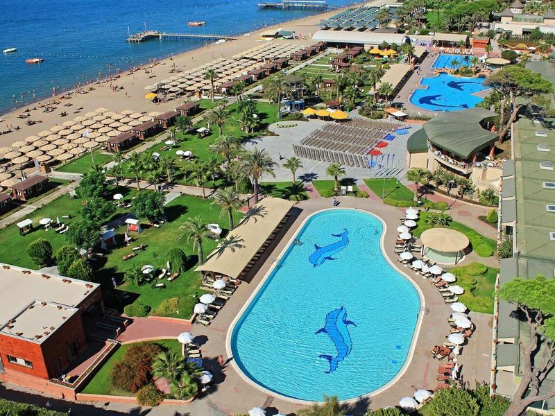 Blick auf Hotelanlage mit großer Pool-Landschaft, Sonnenschirmen, Palmen und Strand am Meer.