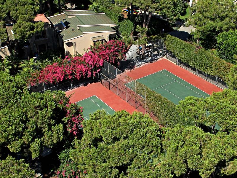 Ein Tennisplatz umgeben von Bäumen und grünen Pflanzen, daneben ein Gebäude mit Dach in hellem Farbton.