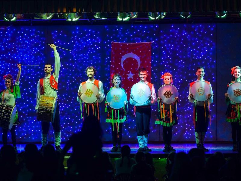 Acht Männer stehen auf Bühne, halten traditionelle Trommeln, türkische Flagge im Hintergrund.