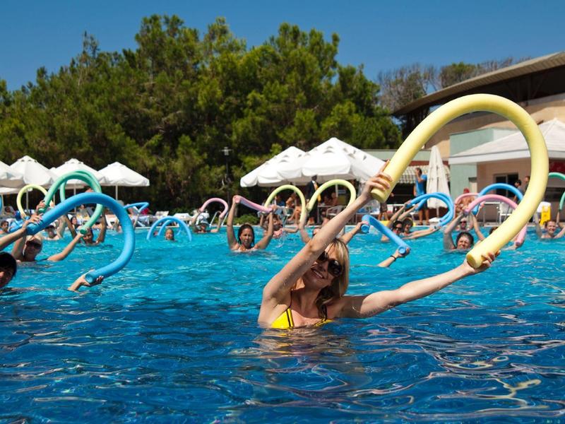 People doing water aerobics with foam noodles in an outdoor pool under sunny skies.