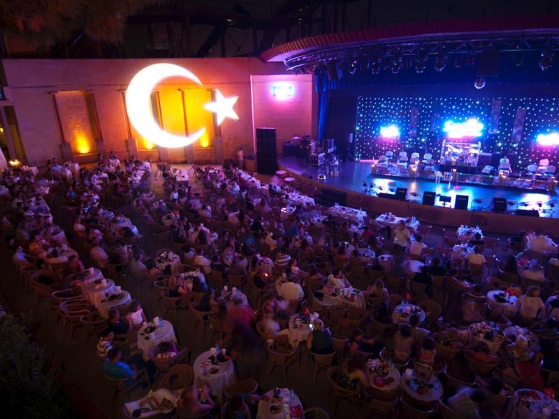 Large crowd at evening event with stage and Turkish flag.