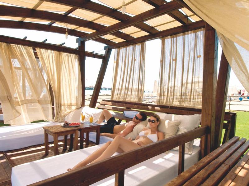Relaxed person on sunbed under wooden pergola with sheer curtains by the water