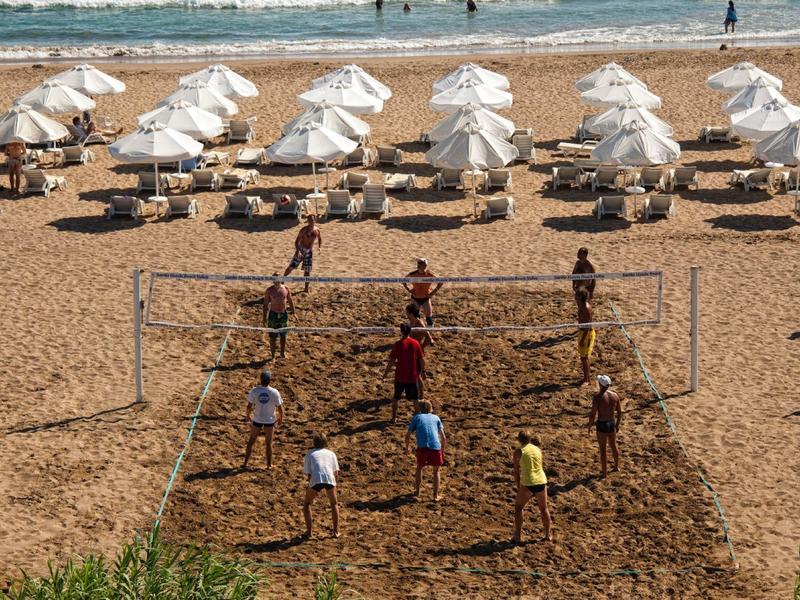 People playing beach volleyball on sandy beach with sun loungers and umbrellas in the background.