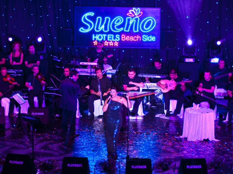 Musicians perform live on stage with the illuminated Sueno Hotels Beach Side logo in the background.