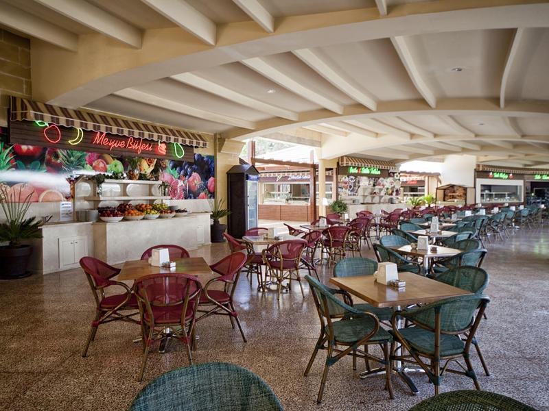 Covered outdoor hotel terrace with tables and chairs, surrounded by plants.