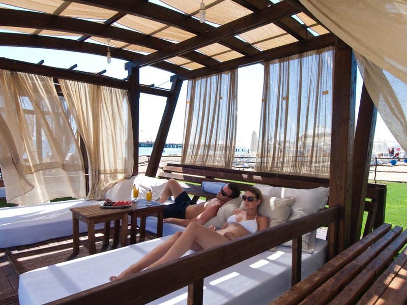 Two people relax on a covered lounge with curtains in the outdoor area of a hotel.