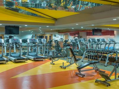 Großer Fitnessraum mit Laufbändern und Kraftgeräten, helle gelbe und orange Akzente im Boden.