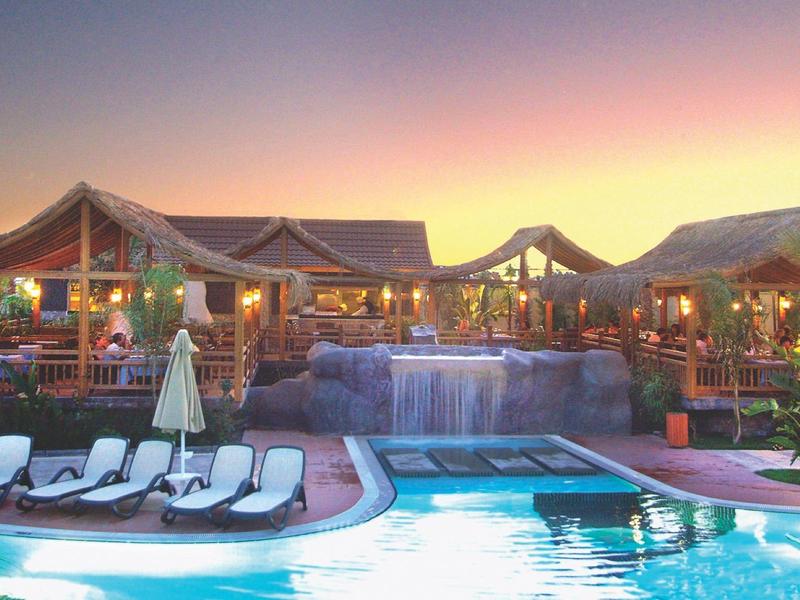 Piscine éclairée au crépuscule avec chaises longues et restaurant en plein air convivial.