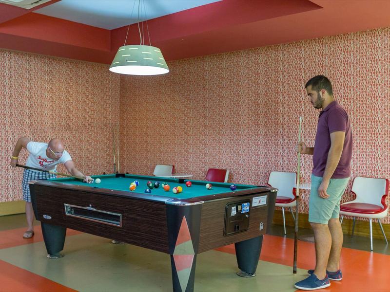 Deux hommes jouent au billard dans une salle avec un mur orange et des chaises blanches.