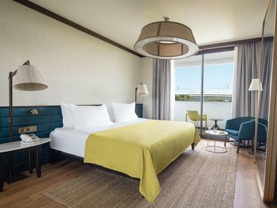 Habitación de hotel moderna con cama grande, manta amarilla, dos mesas y vista al mar por la ventana.