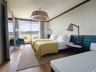 Habitación de hotel luminosa con cama doble, sillones, escritorio y vista al lago a través de la ventana.