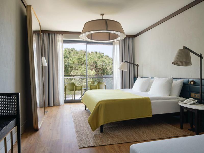 Habitación de hotel con cama doble y ventana grande con vista a árboles y balcón.