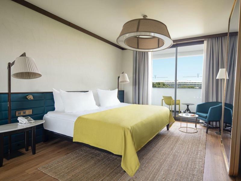 Habitación de hotel moderna con cama grande, manta amarilla, dos mesas y vista al mar por la ventana.