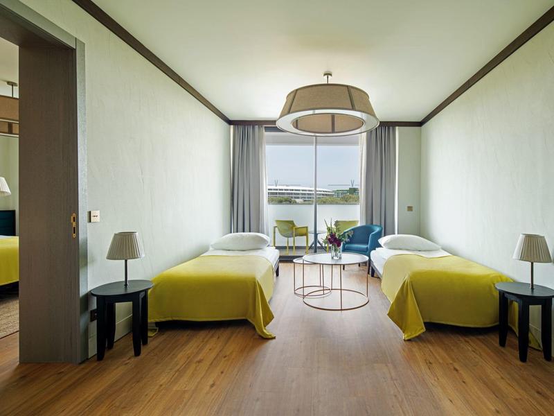 Habitación de hotel luminosa con dos camas individuales y vista al balcón y al agua.