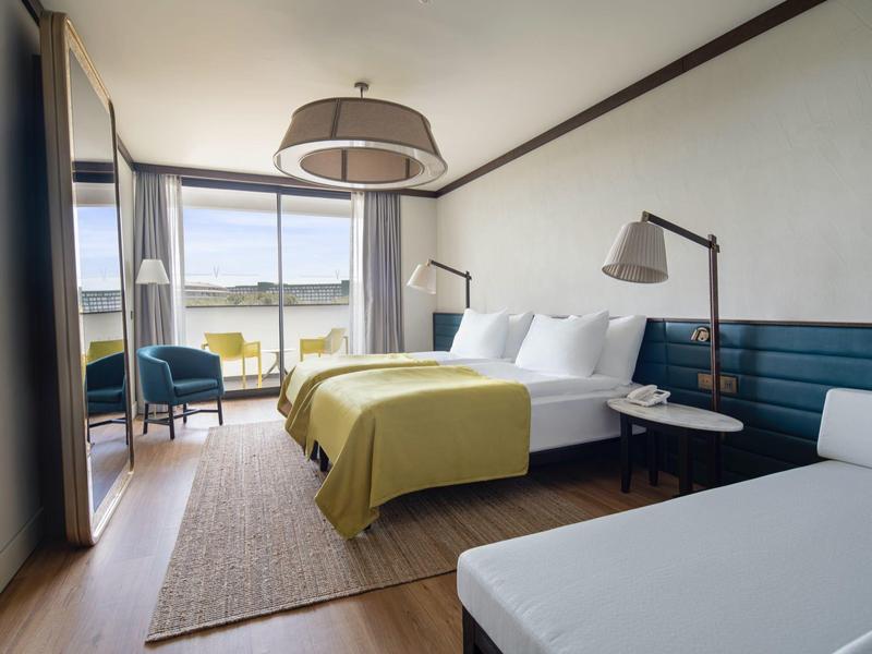 Habitación de hotel luminosa con cama doble, sillones, escritorio y vista al lago a través de la ventana.