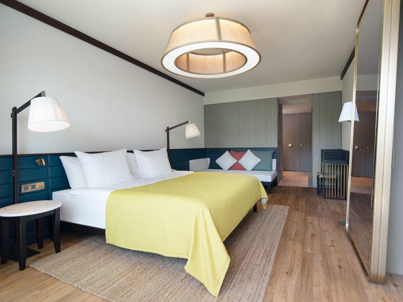 Habitación de hotel moderna con cama doble grande, colcha amarilla, suelo de madera y lámparas elegantes.