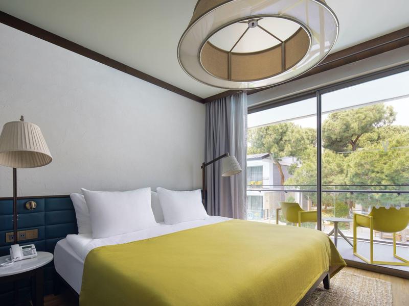 Habitación de hotel moderna con cama grande, cubrecama verde y balcón con sillas y vista a árboles.