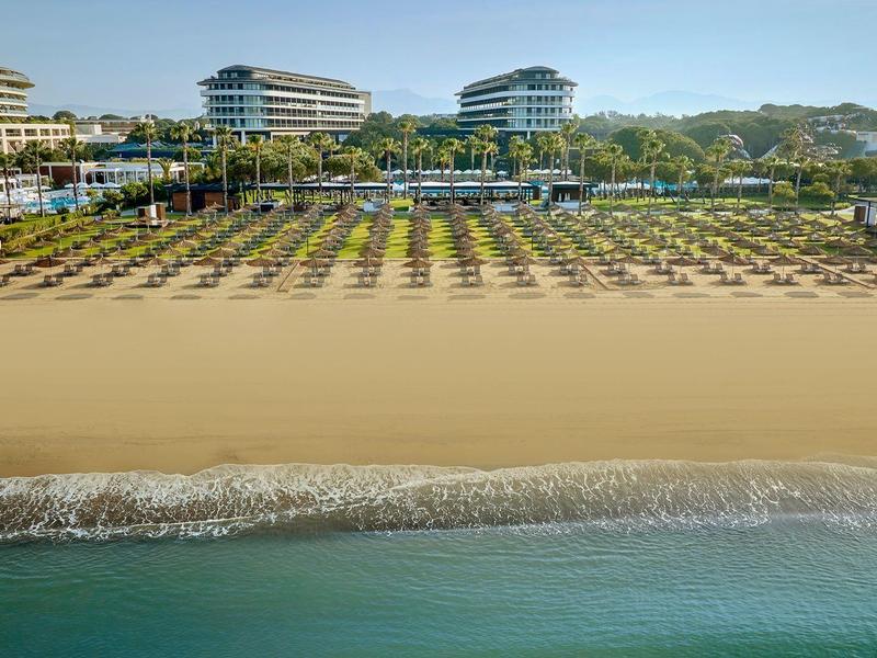 Vista de una amplia playa de arena con un resort hotelero y filas de sombrillas al fondo.