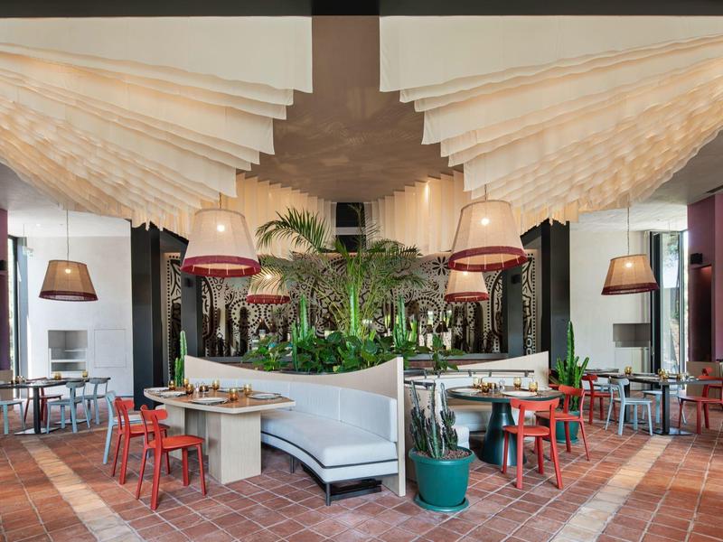 Restaurante moderno de hotel con plantas centrales, sillas de colores y lámparas colgantes.