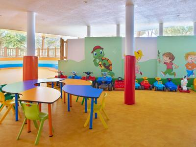Bunter Kindergartenraum mit Tischen, Stühlen und Wandmalerei von spielenden Kindern.