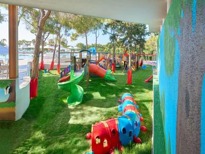 Bunter Spielplatz mit Rutschen, Klettergeräten und grünem Gras unter Bäumen am Wasser.